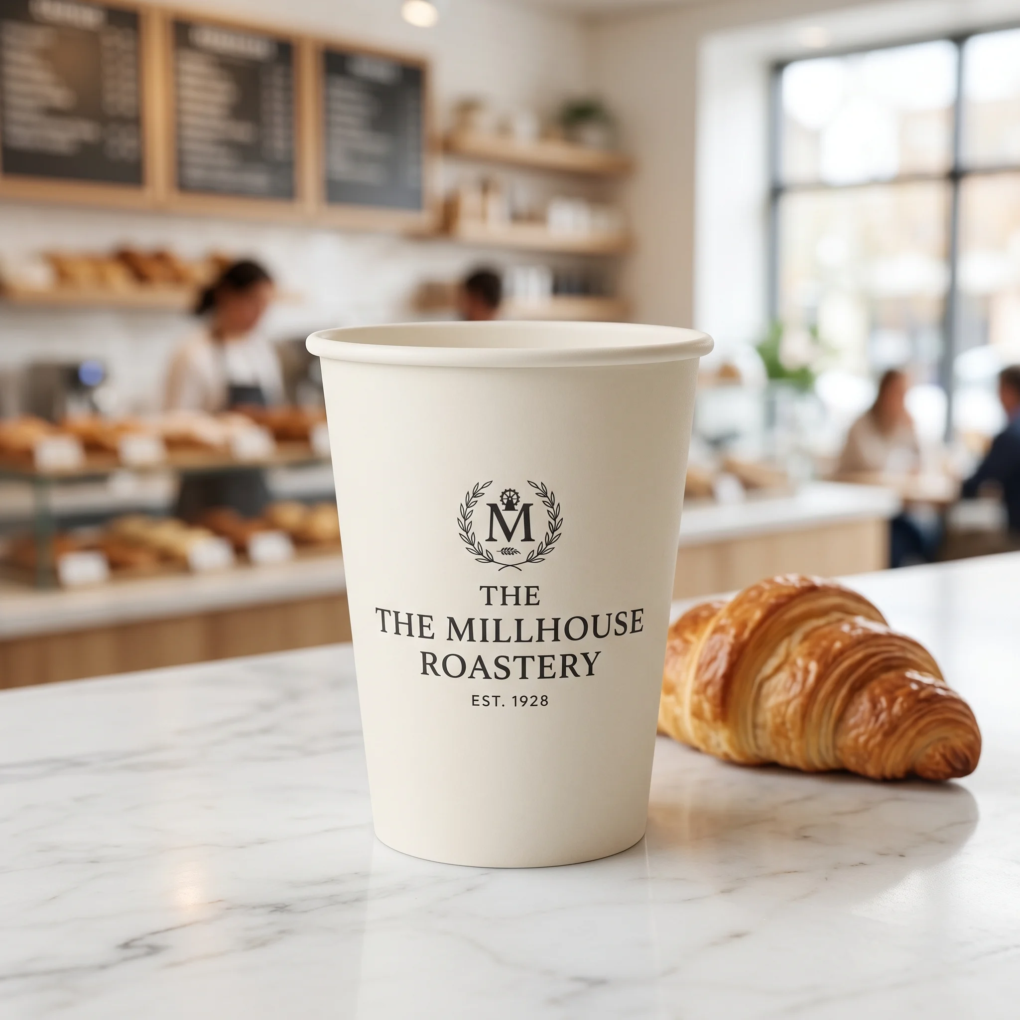 White coffee cup with The Millhouse Roastery logo next to golden croissant on marble counter in bright modern bakery
