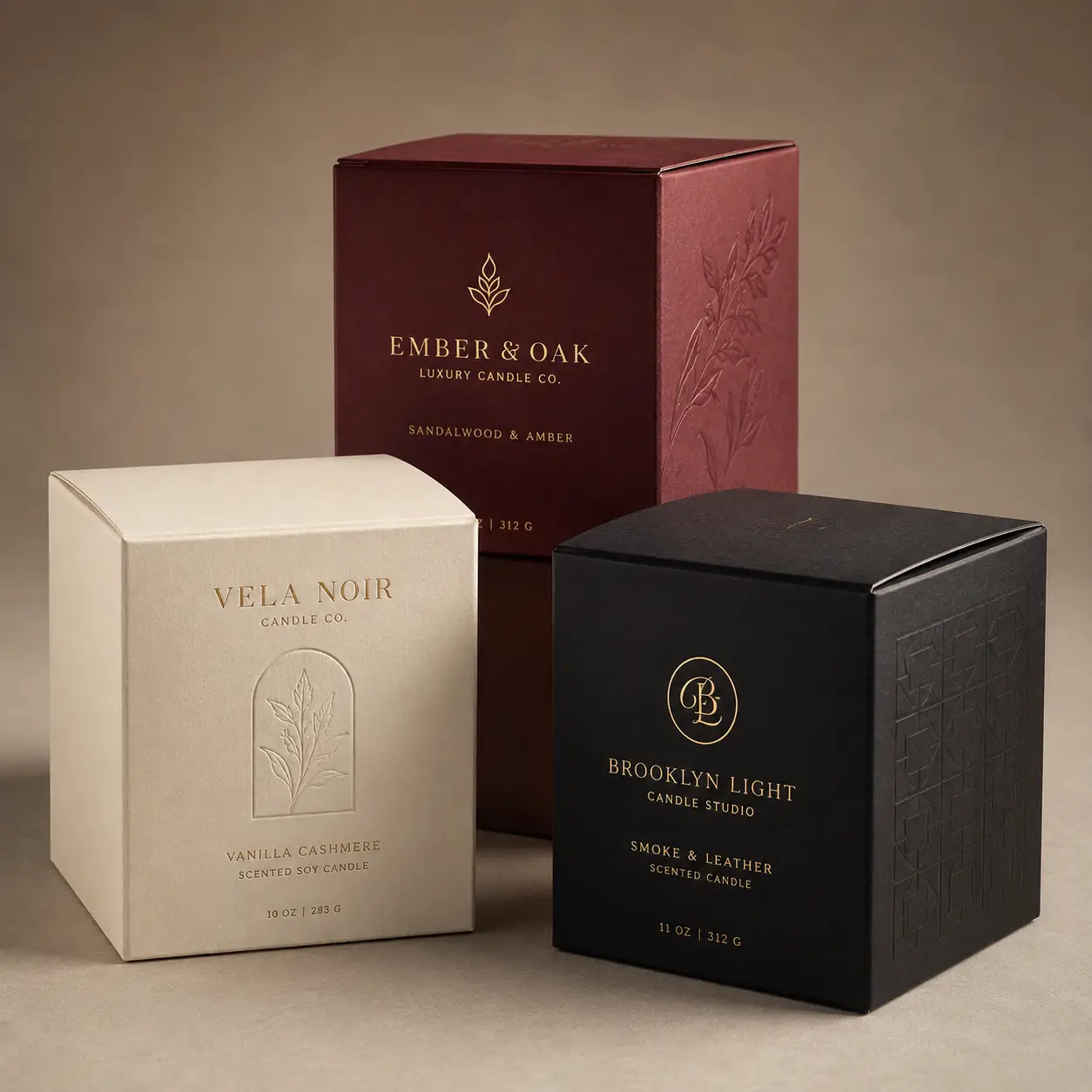 Three luxury candle boxes in cream, black, and burgundy with gold foil branding and embossed floral designs on neutral background