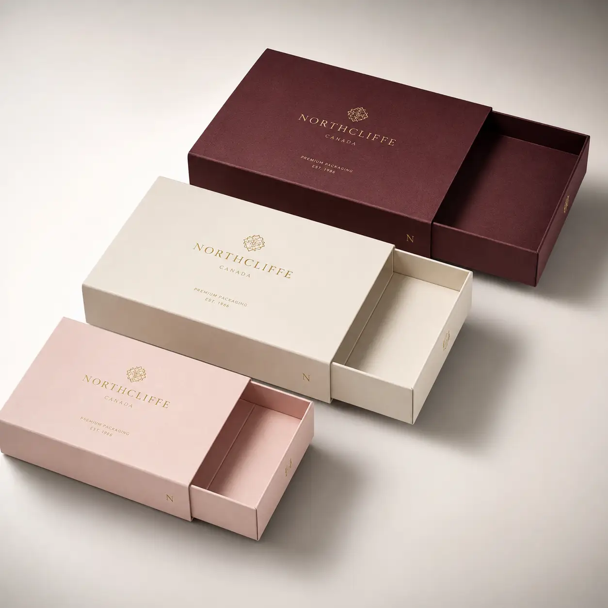 Three luxury slide drawer gift boxes in burgundy, cream, and blush pink with gold Northcliffe branding, premium packaging design