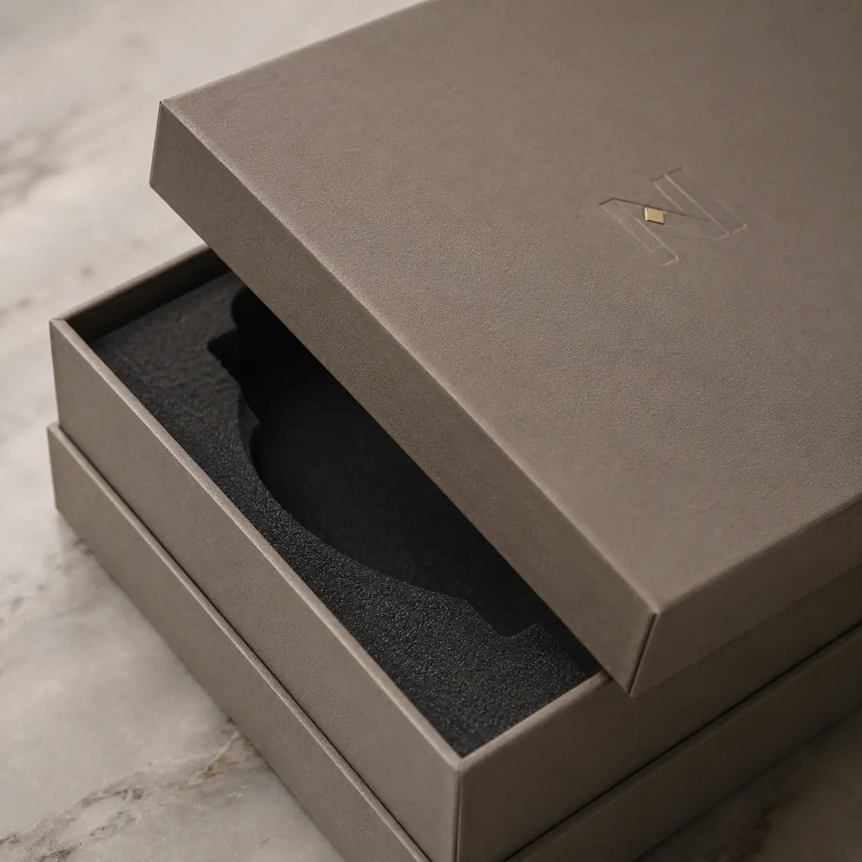 Luxury taupe watch gift box with foam insert and gold embossed logo, opened on marble surface, premium packaging design