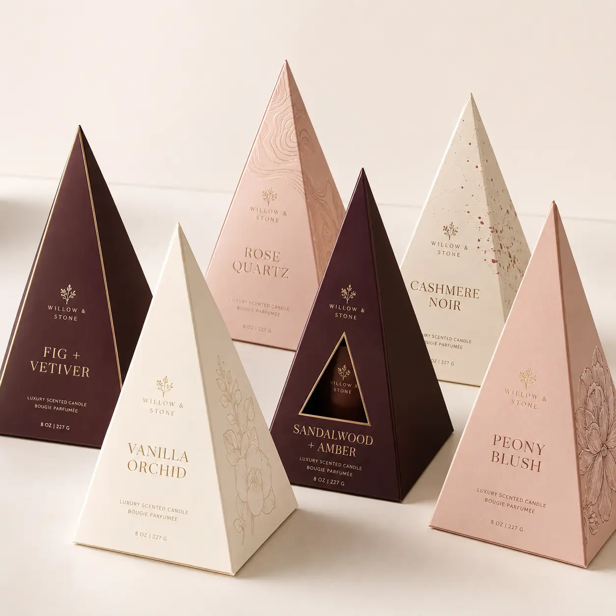 Six luxury pyramid candle boxes by Willow & Stone in blush, cream, and burgundy with gold foil scent labels on white surface
