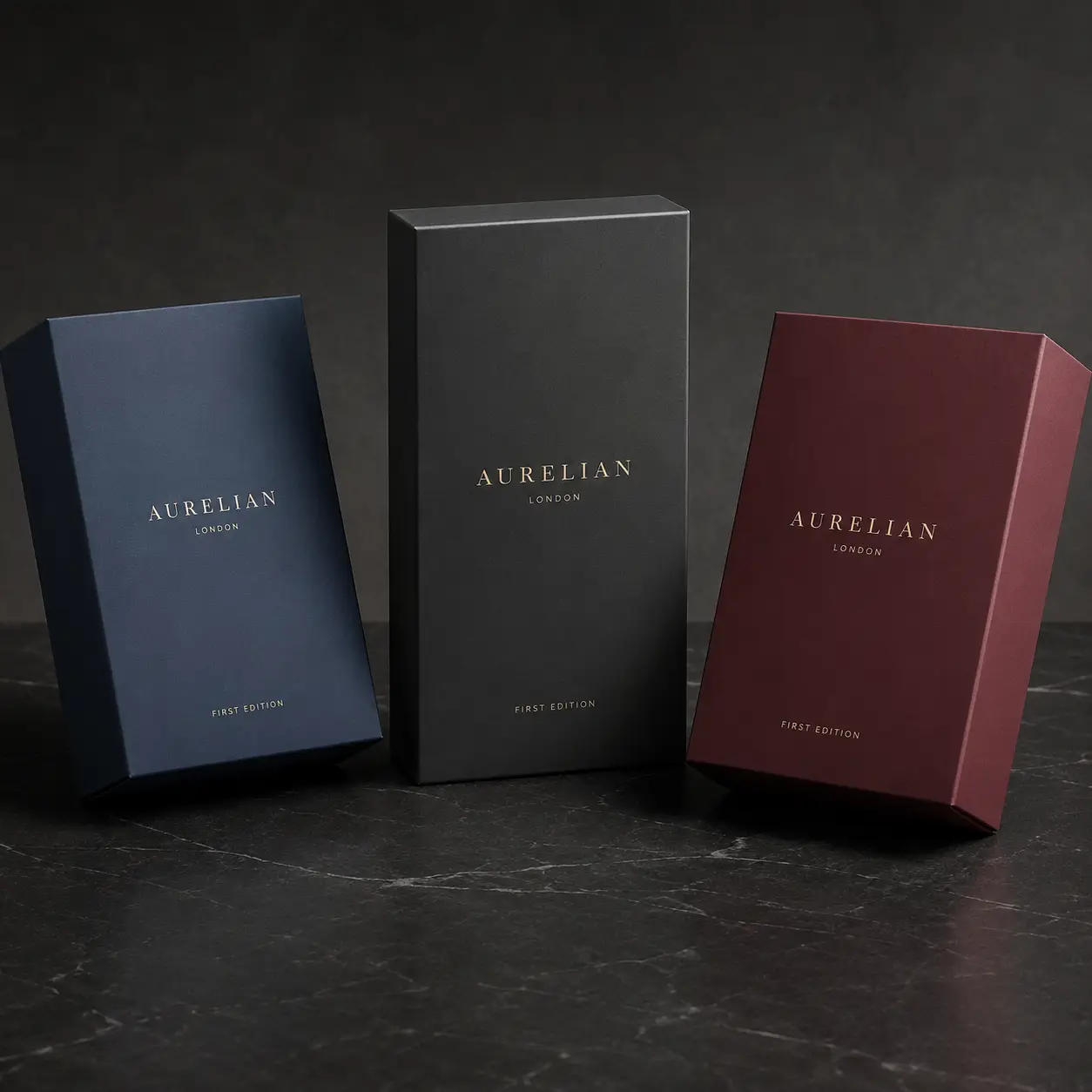 Three luxury Aurelian London First Edition gift boxes in navy, charcoal, and burgundy with gold foil branding on dark marble