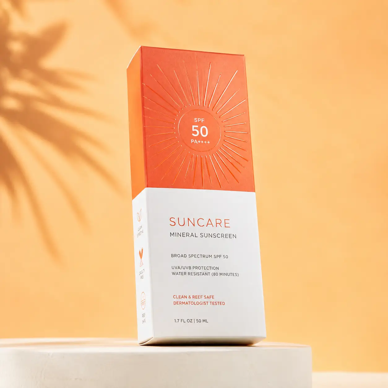 Orange and white SPF 50 mineral sunscreen packaging box on white pedestal, warm golden background with palm shadow, reef-safe formula