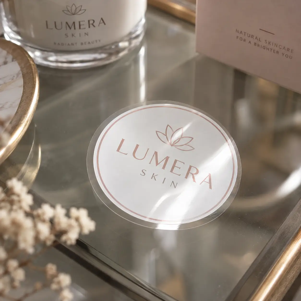 Lumera Skin rose gold logo sticker on mirrored tray beside white cream jar and pink card, elegant custom packaging branding display