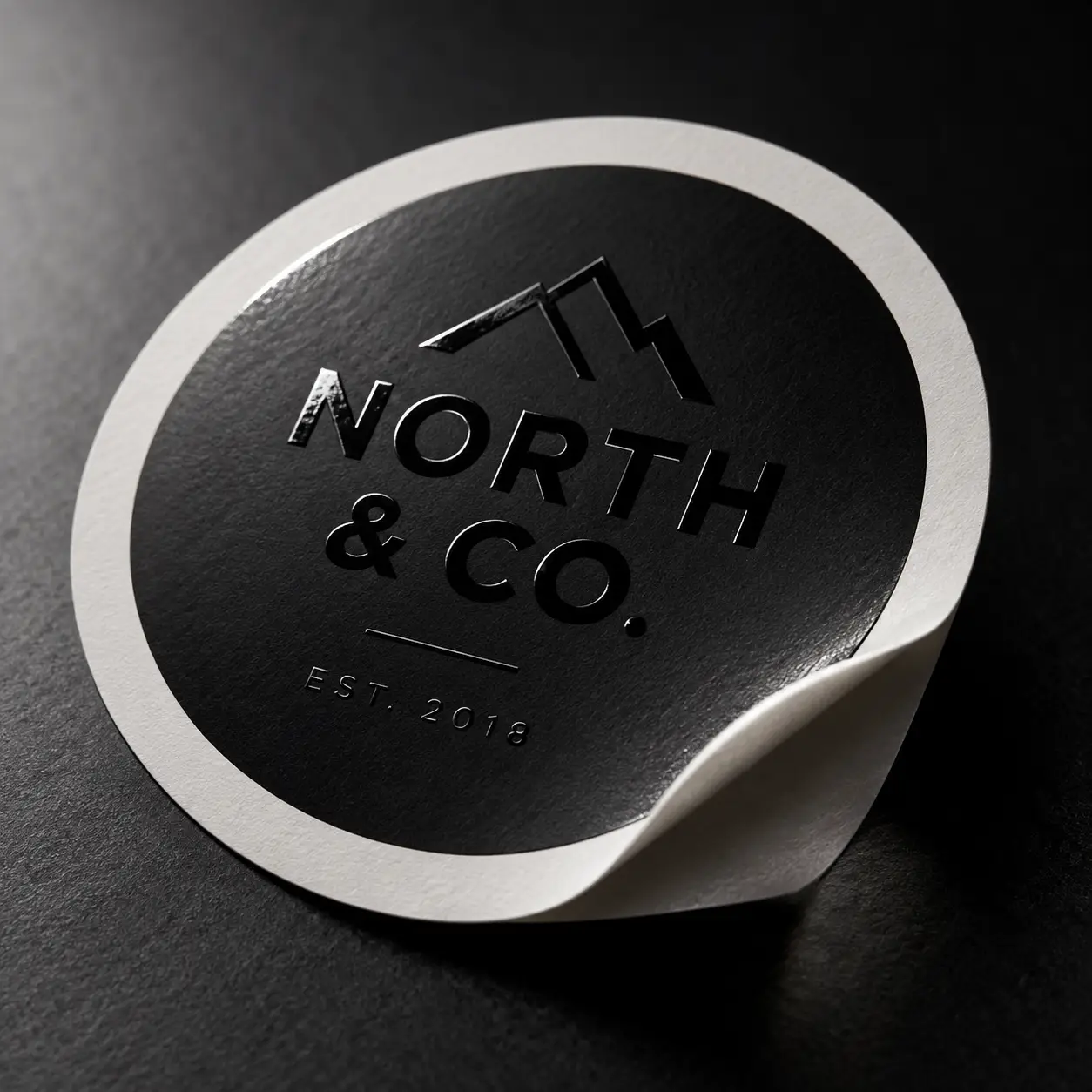 Black round sticker label with embossed North & Co. Est. 2018 logo, mountain graphic, peeling corner, dark moody product packaging mockup
