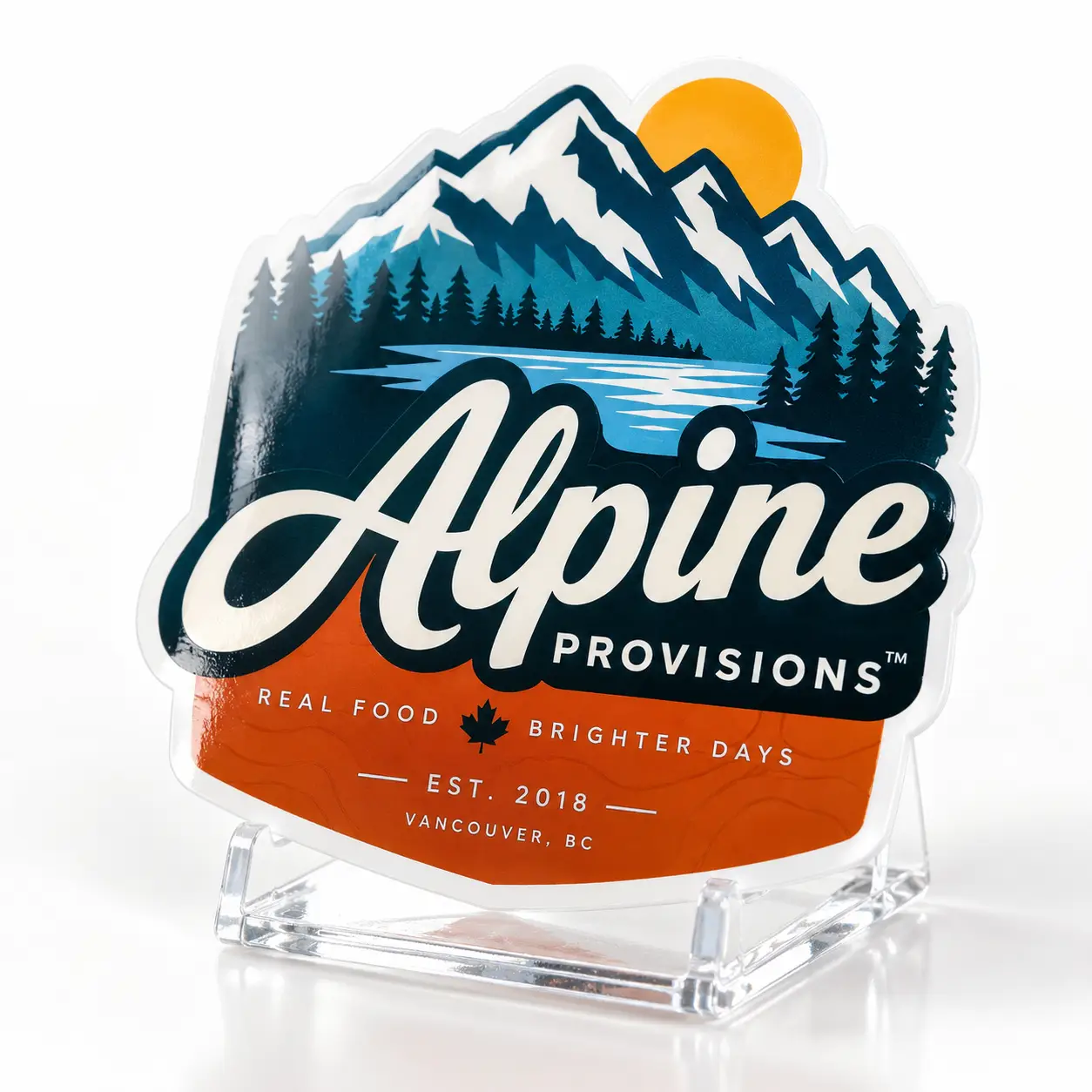 Alpine Provisions die-cut sticker with mountain lake scene, orange base, navy script logo, displayed on clear acrylic stand