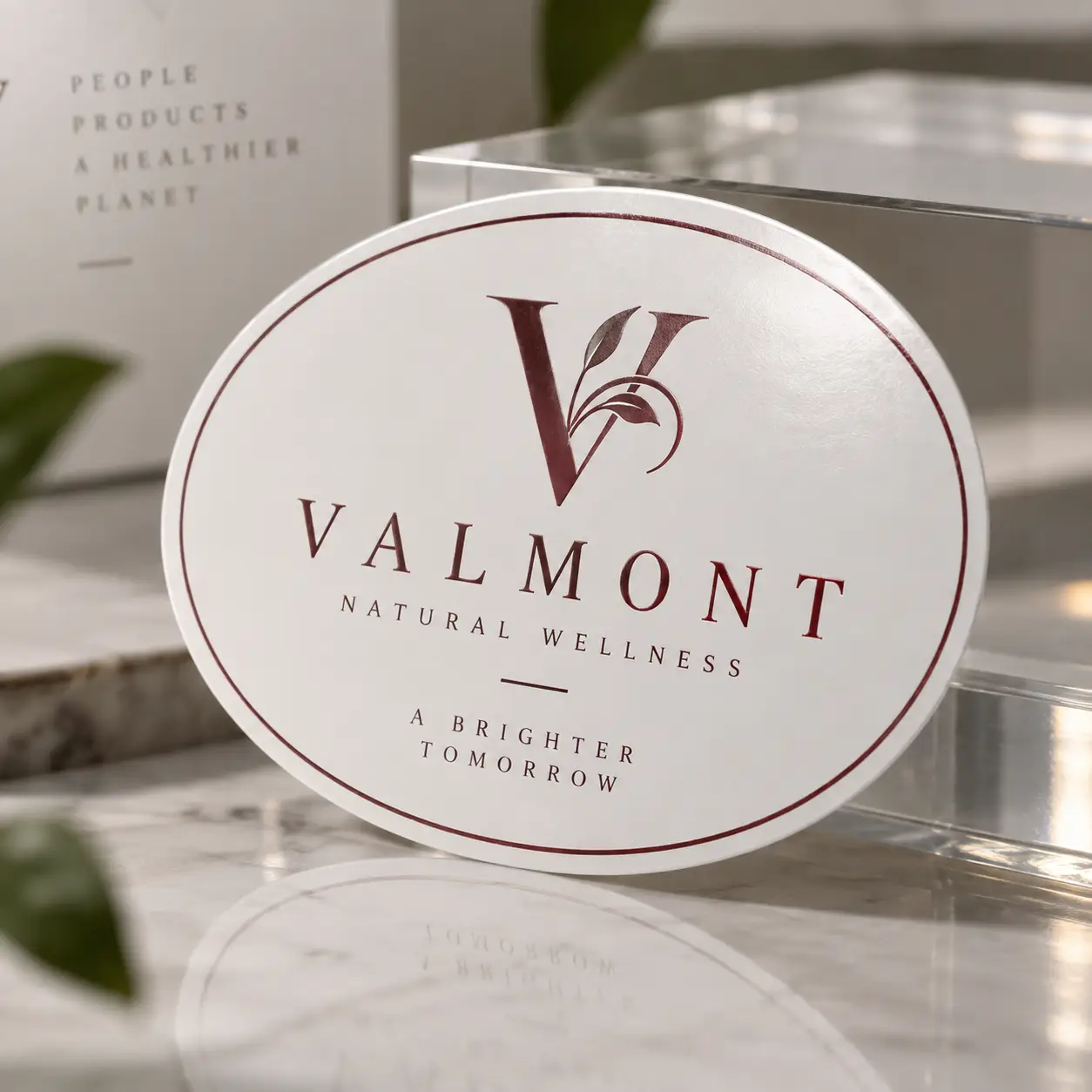 Elegant oval white sticker label for Valmont Natural Wellness brand in burgundy, displayed on marble surface with acrylic block