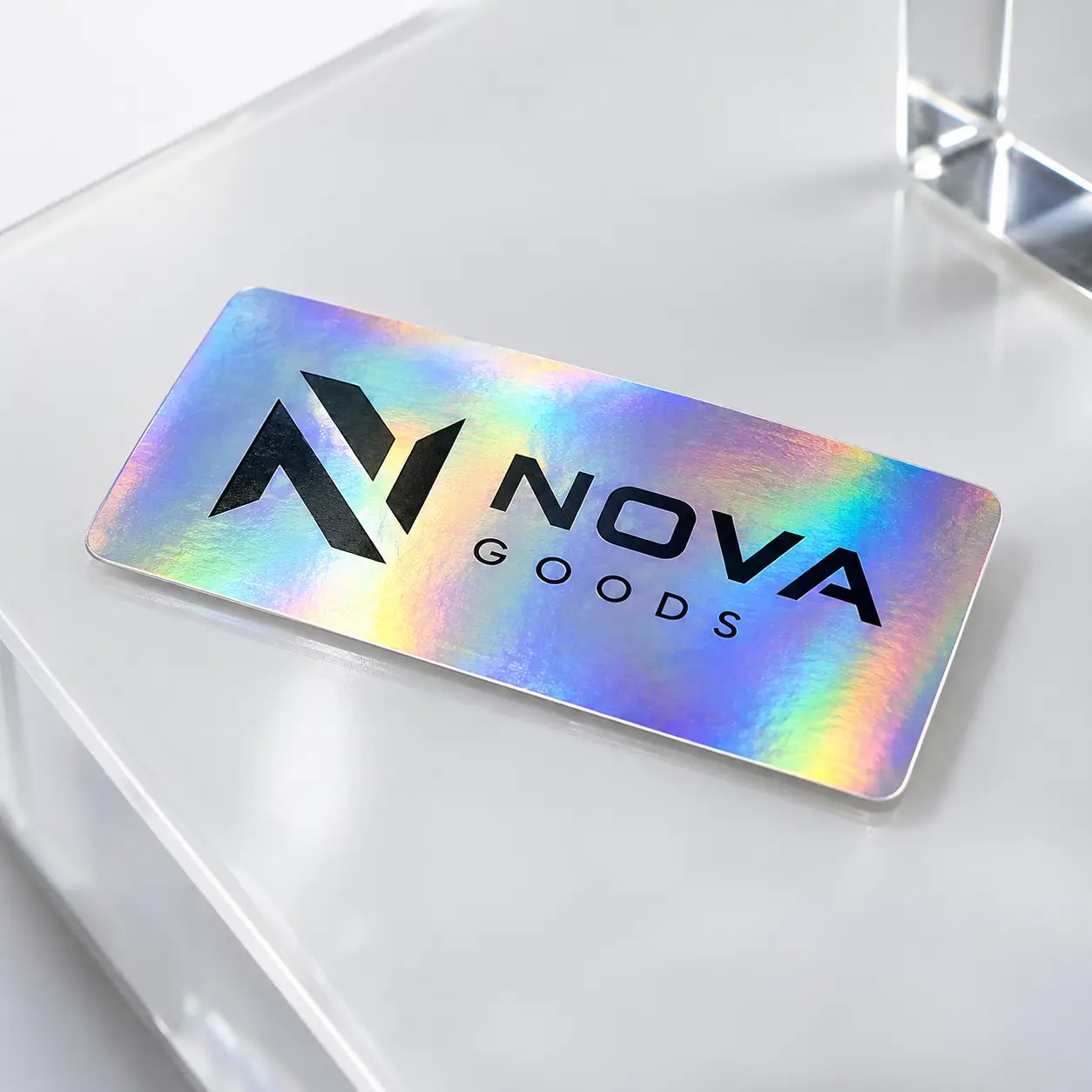 Holographic sticker label with Nova Goods logo in black on iridescent rainbow foil, displayed on white acrylic surface