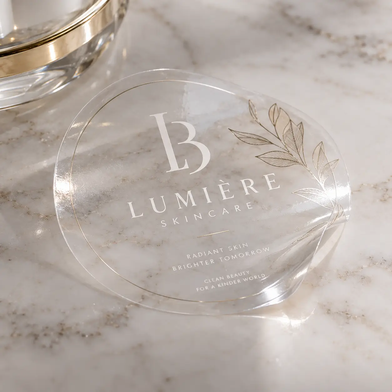 Lumière Skincare clear acrylic round logo coaster with gold botanical design on white marble surface