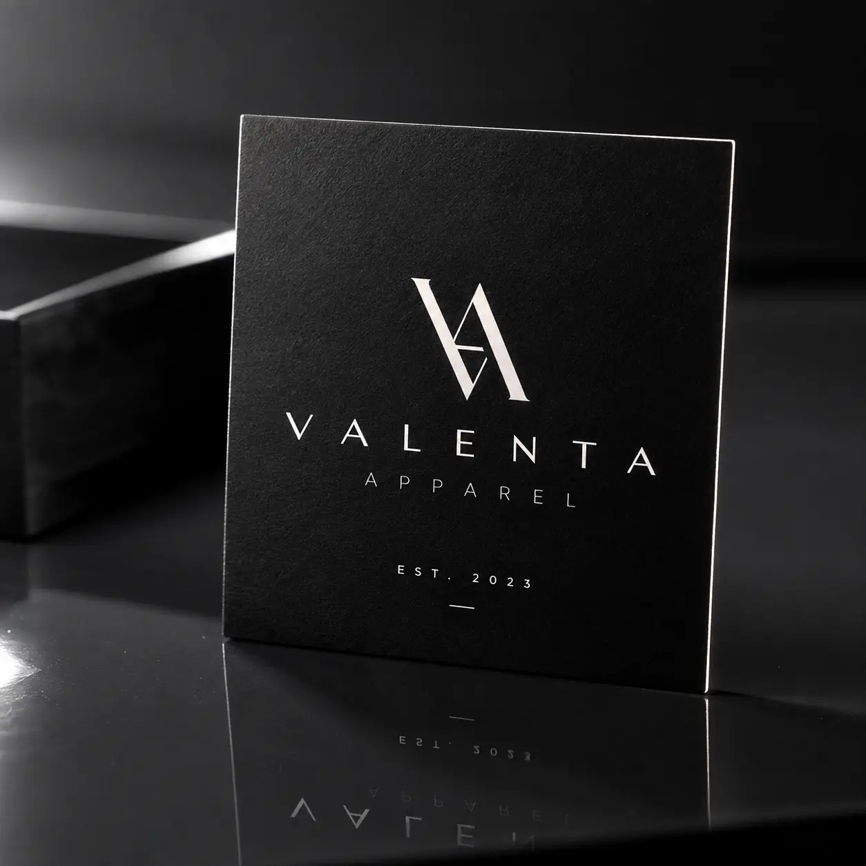 Luxury black matte business card for Valenta Apparel Est. 2023 with silver foil logo, leaning against black box on reflective surface