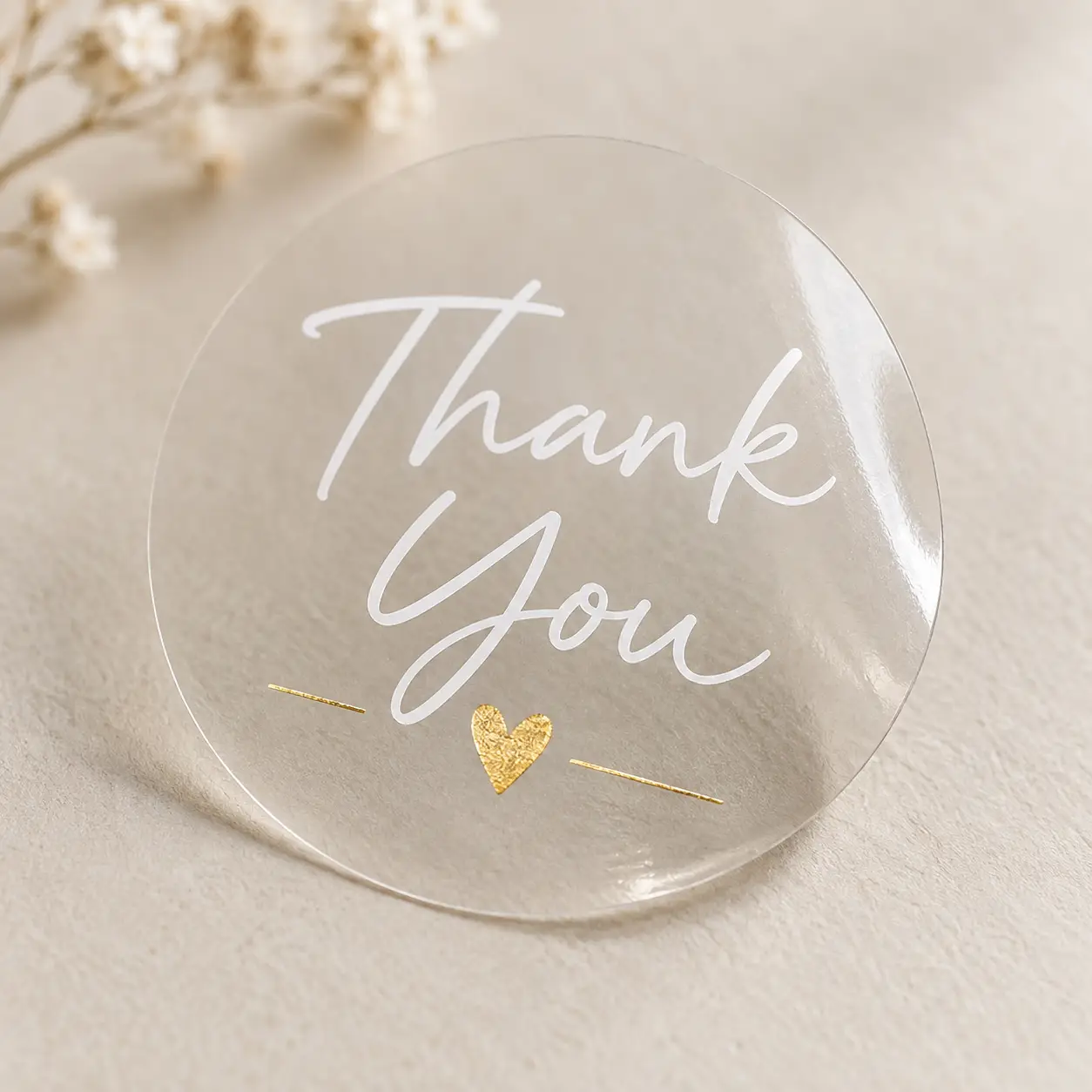Clear round Thank You sticker with white script text and gold foil heart, displayed on beige surface with white flowers