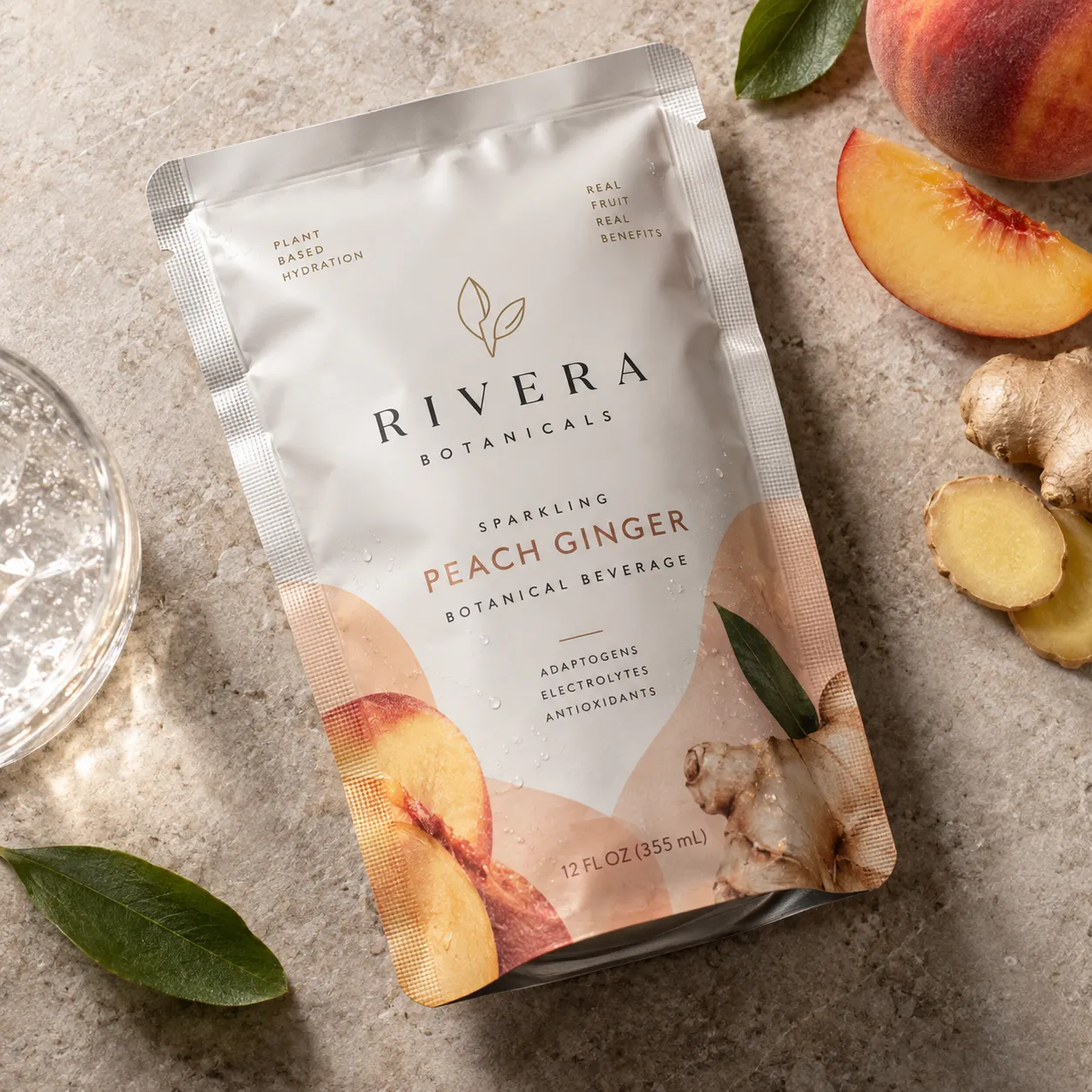 Rivera Botanicals sparkling peach ginger botanical beverage pouch, 12oz, surrounded by fresh peaches and ginger root on stone surface