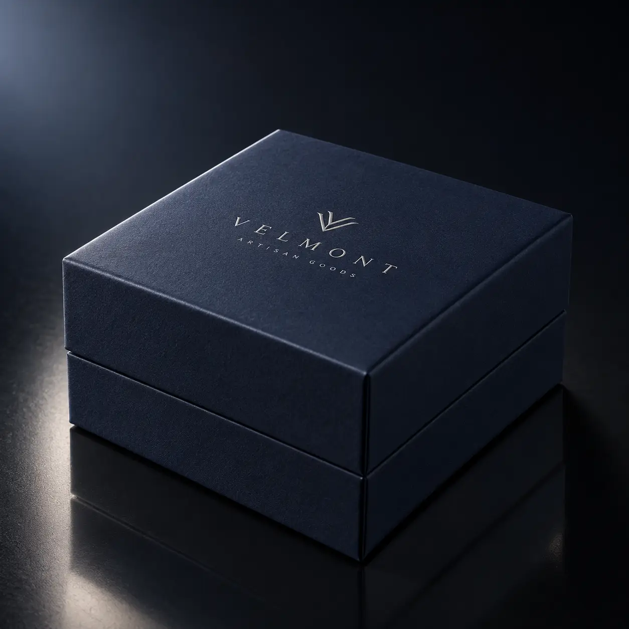 Velmont Artisan Goods navy blue luxury gift box with silver foil logo on dark reflective surface, premium packaging design